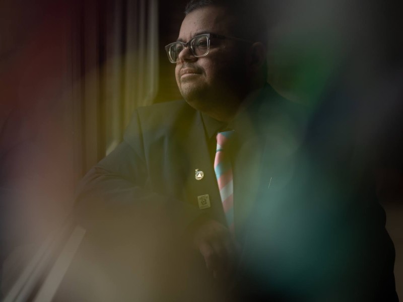 Aim Comperatore wears a black suit and a tie with the stripes of the transgender pride flag while posing for a portrait at Comperatore's Pressley Street High Rise apartment.