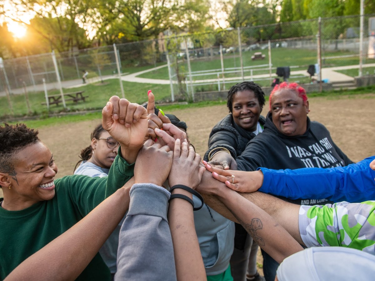 From left, Kia Foster, of Penn Hills, Jasmine “Happy Feet” Thomas, of East Liberty, team manager Yvette Harrison, of Wilkinsburg, Sandra “Ma Duke” Douglass, of the Hill District, and coach Haywood “Cheddar Melt” El, of Wilkinsburg, come together at the end of practice for their Wilkinsburg Alive and Kicking Kickball Team on April 26 at Whitney Park in Wilkinsburg. Games start on Sundays in June, and are broadcast on YouTube. (Photo by Stephanie Strasburg/PublicSource)