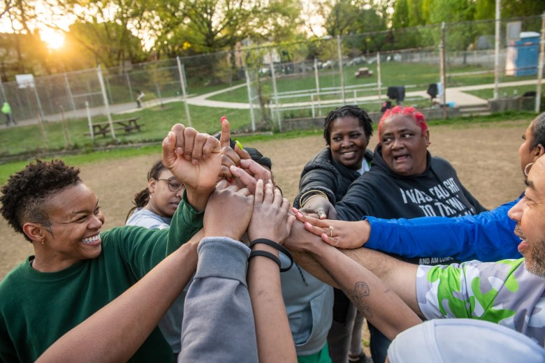 From left, Kia Foster, of Penn Hills, Jasmine “Happy Feet” Thomas, of East Liberty, team manager Yvette Harrison, of Wilkinsburg, Sandra “Ma Duke” Douglass, of the Hill District, and coach Haywood “Cheddar Melt” El, of Wilkinsburg, come together at the end of practice for their Wilkinsburg Alive and Kicking Kickball Team on April 26 at Whitney Park in Wilkinsburg. Games start on Sundays in June, and are broadcast on YouTube. (Photo by Stephanie Strasburg/PublicSource)