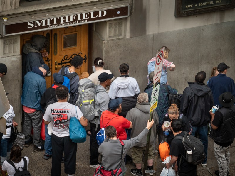 People wait to get in the Smithfield United Church of Christ shelter in the minutes before it opens for the night on May 8, 2023, in downtown Pittsburgh. Above the wooden door, a sign reads "Smithfield." Beside, a sign reads "HAVE A BEAUTIFUL DAY." Underneath, people hold their bags and gather tightly to get their spot in the shelter. (Photo by Stephanie Strasburg/PublicSource)