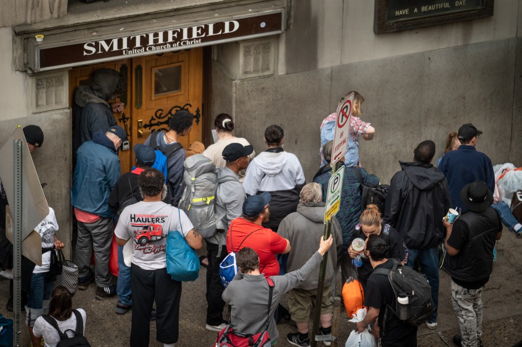 People wait to get in the Smithfield United Church of Christ shelter in the minutes before it opens for the night on May 8, 2023, in downtown Pittsburgh. Above the wooden door, a sign reads "Smithfield." Beside, a sign reads "HAVE A BEAUTIFUL DAY." Underneath, people hold their bags and gather tightly to get their spot in the shelter. (Photo by Stephanie Strasburg/PublicSource)