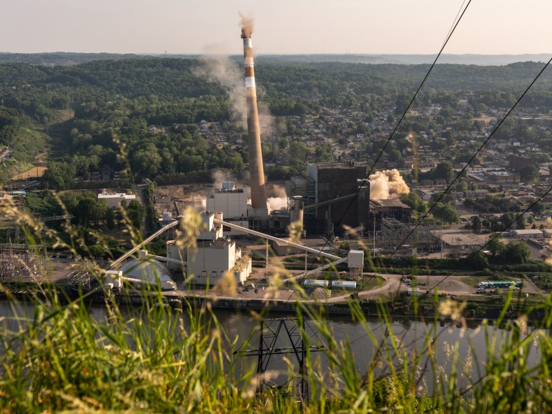 The smokestacks at the former Cheswick Generating Station in Springdale fall to the ground during the implosion of the stacks on Friday, June 2, 2023, as seen from Barking Slopes Conversation Area across the Allegheny River. (Photo by Stephanie Strasburg/PublicSource)