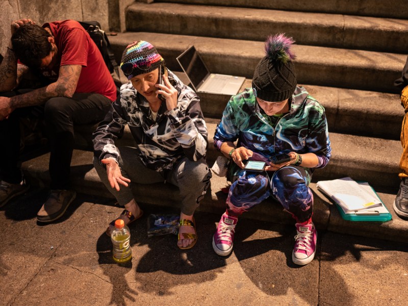 In her signature crocheted rainbow hat and smoke printed sweats, Aubrey Plesh, center, left, founder of Team PSBG, calls area shelters to secure other housing for former guests of the Smithfield United Church of Christ homeless shelter as she sits on the church steps on the evening of Wednesday, June 21, 2023, in Pittsburgh’s downtown. Plesh and PSBG team member Amanda Fry, right, were trying to coordinate how people would get to other shelter beds after the controversial space closed that morning at 7 a.m. At left and right, people in need of alternative shelter sit on the church steps waiting for a placement. (Photo by Stephanie Strasburg/PublicSource)