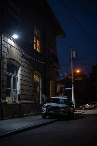 A blue glow from an overhead light casts shadows across a Pittsburgh Police vehicle parked along a sidewalk outside a police station at night. A window above glows yellow. A streetlight casts a dim light in the background. (Photo by Stephanie Strasburg/PublicSource)