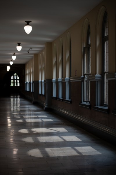 Light and shadows make a pattern across the Allegheny County Courthouse floor, where the district attorney’s office is. The perspective leads to a window in darkness at the very back. Above, hallway lights hang in even increments along the ceiling. (Photo by Stephanie Strasburg/PublicSource)