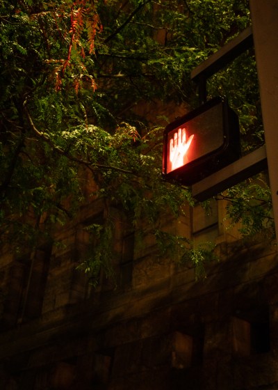 A red hand traffic signal glows at night outside the Allegheny County Courthouse, where the district attorney’s office is, on Aug. 16, 2023, in Pittsburgh. Leaves reflect the red glow of the signal and the yellow cast of a street light. Behind, the stone facade of the courthouse fades to black. (Photo by Stephanie Strasburg/PublicSource)