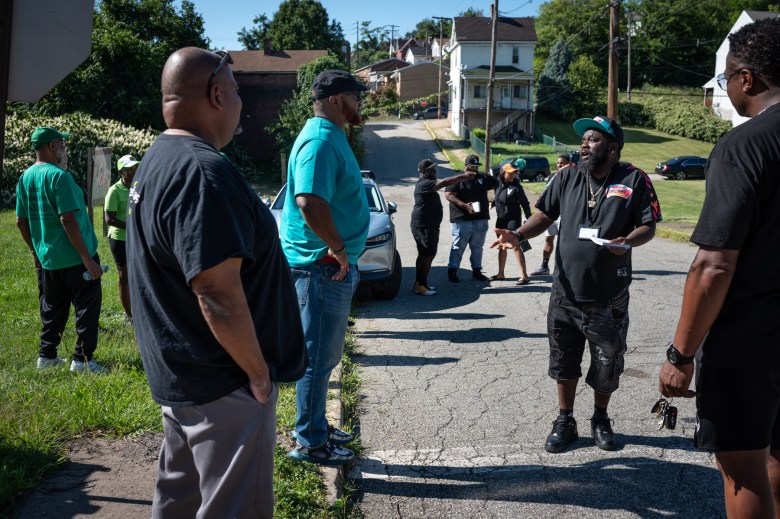 Lee Davis, third from left, director of violence prevention for Greater Valley Community Services (GVCS), listens to Kenneth Woods, second from right, a violence interrupter with GVCS, as they gather with other outreach teams ahead of a vigil for two teens fatally shot several nights prior in the surrounding grassy lots, on Thursday, Aug. 31, 2023, in Braddock. Woods waved informational pamphlets for trauma support and mentorship opportunities as rush hour traffic and school busses rolled by the scene. “We out in the community every day so when they see us they already know what we’re doing out here,” said Woods. “It’s basically based on our past relationships inside of the community.” (Photo by Stephanie Strasburg/PublicSource)