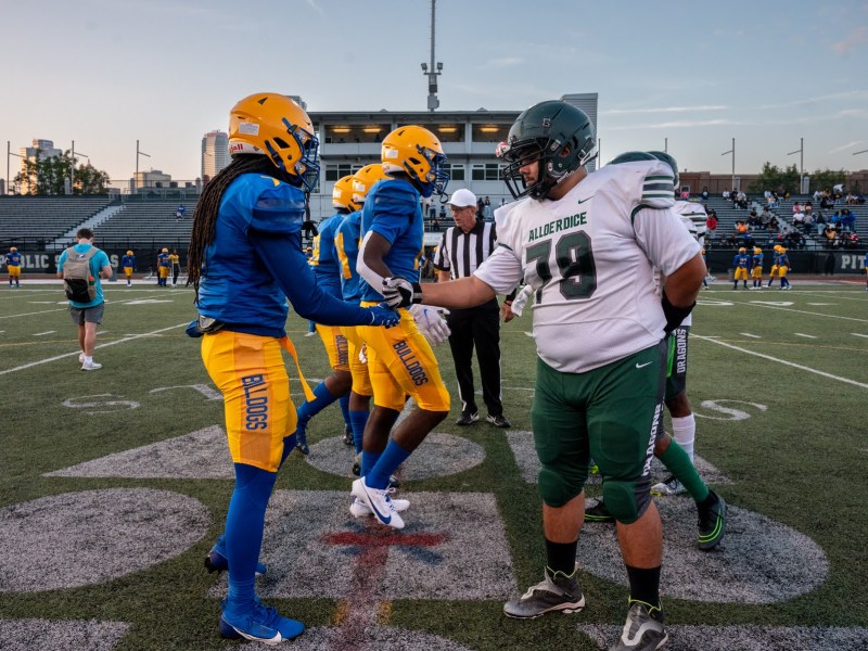 The Pittsburgh Westinghouse Academy 6-12 Bulldogs, left, shake hands with the Taylor Allderdice High School Dragons at the start of their football game, Thursday, Sept. 21, 2023, at Cupples Stadium in the South Side. The high schools sit only three miles apart but the disparities range from academic programming to infrastructure. At left, the blue and yellow uniformed Bulldogs, at right, white and green Dragons. The two players at front pound fists as a greeting. (Photo by Stephanie Strasburg/PublicSource)