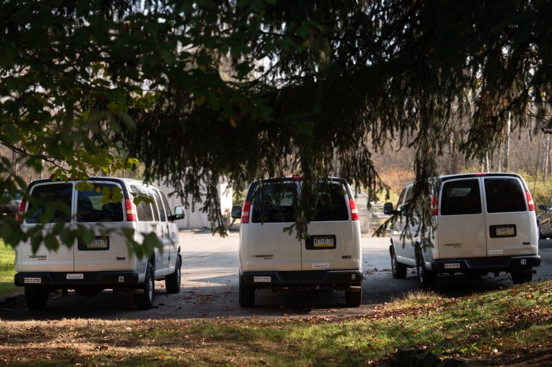 White transport vans are parked at Adelphoi Village. They can be seen beyond the low-hanging branches of trees.  