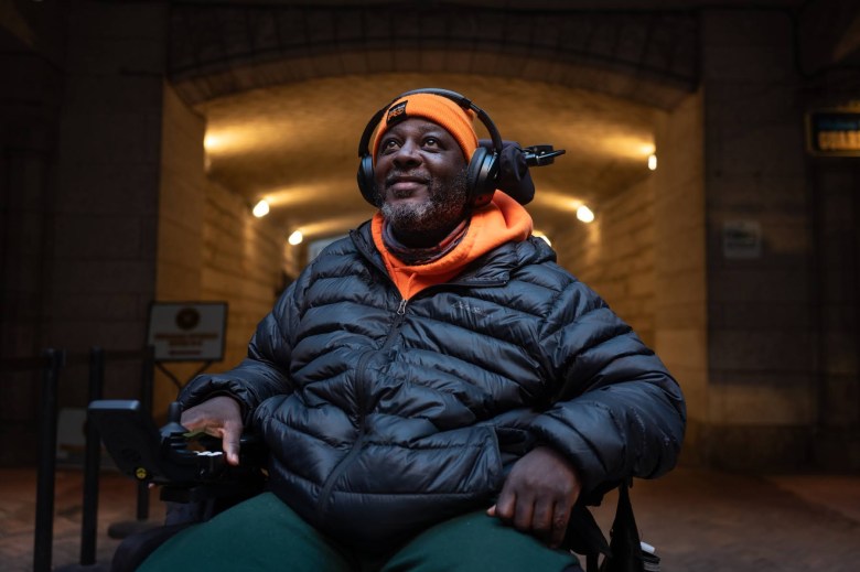 Walter Harris is posing for a portrait outside the Allegheny County Courthouse. He's in a wheelchair and is wearing an orange ski cap, a black puffer jacket and green sweatpants.