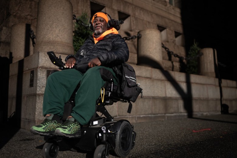 Walter Harris, who's in a wheelchair, poses for a portrait outside the Allegheny County Courhouse. He's wearing an orange ski cap, a black puffer jacket and green sweatpants. 