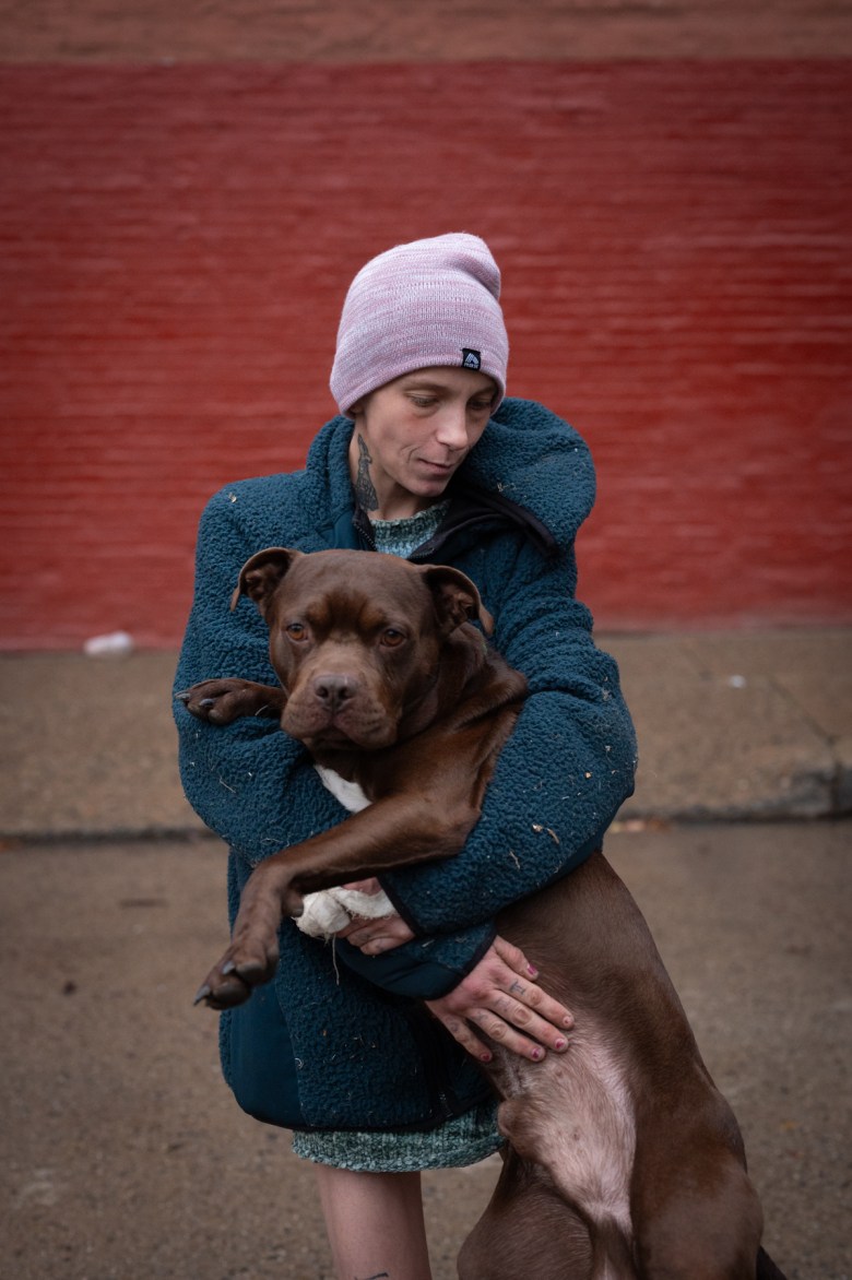 A person in a winter hat and fleece living at the First Ave. encampment holds her brown dog for a portrait near to her tent, on Sunday, Oct. 29, 2023, in downtown Pittsburgh. She says she misses the community at the winter shelter at the Smithfield United Church of Christ. (Photo by Stephanie Strasburg/PublicSource)