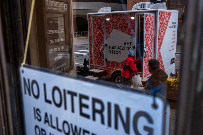 One of two portable public restrooms that were added in the ongoing conflict about homelessness in downtown Pittsburgh is reflected in the glass doors above the Smithfield United Church of Christ steps, on Friday, Nov. 3, 2023, in downtown Pittsburgh. The church, which held a winter emergency shelter in the basement for decades that became controversial as the need for the space stretched past its normal winter months earlier this year, continues to be a gathering point for people. (Photo by Stephanie Strasburg/PublicSource)