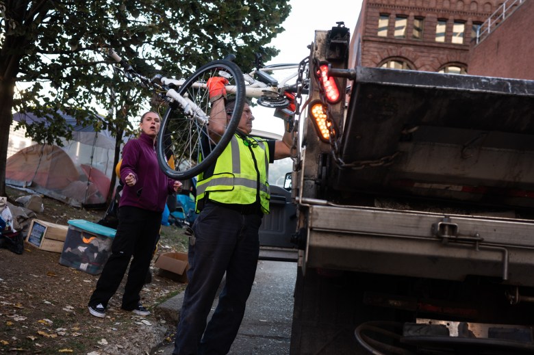 A Department of Public Works employee loads a truck with the belongings of a person staying at the First Avenue encampment for storage, on Tuesday, Nov. 7, 2023, in downtown Pittsburgh. The city’s new encampment policy, finalized in the late summer, states the city is to store people’s belongings for a minimum of 90 days, down from a year in the prior policy. At left, an outreach member watches. (Photo by Stephanie Strasburg/PublicSource)