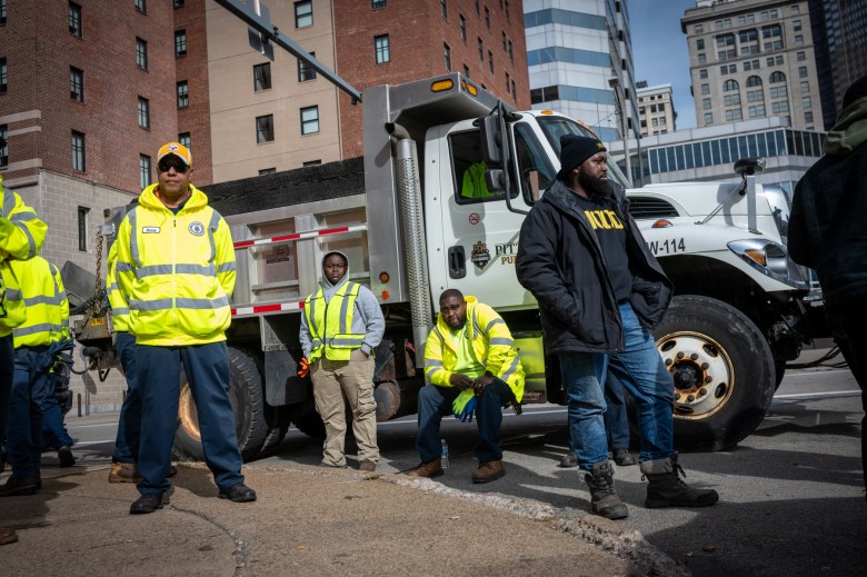 Department of Public Works employees wait with a dump truck to clean up at the site of an encampment along First Avenue by Boulevard of the Allies, on Wednesday morning, Nov. 8, 2023, in downtown Pittsburgh. At right is Muhammad Ali Nasir, also known by his emcee name MAN-E, founder of Community Care & Resistance In Pittsburgh (CCRIP) with 1HOOD, who was present throughout the process to provide supplies, food, and transportation for people living in the camp. “We’re getting requests for sleeping bags, hats, coats, and folks are preparing to sleep outside during the colder months,” he said. (Photo by Stephanie Strasburg/PublicSource)