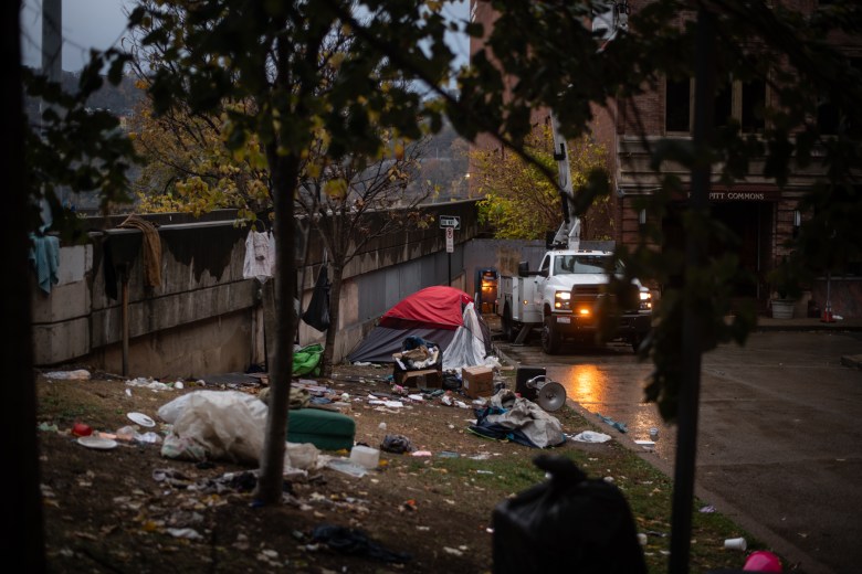 Workers install spotlights on the office building bordering the First Avenue encampment as the wait continues for the Department of Public Works to clear the camp, on Wednesday, Nov. 8, 2023, in downtown Pittsburgh. A flap from the last standing tent blew in the wind as the morning’s clean up was pushed from 7 to 9 to 11 a.m. (Photo by Stephanie Strasburg/PublicSource)