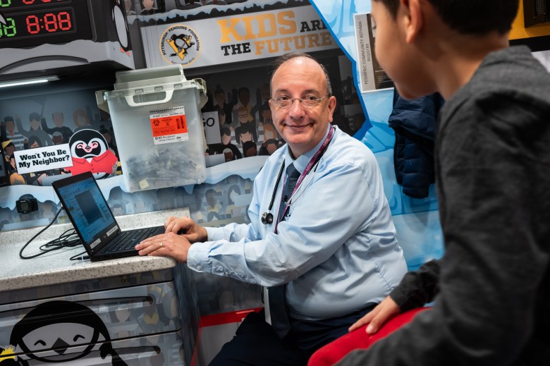 A doctor sitting at a desk with a laptop in front of him talks with a young person.