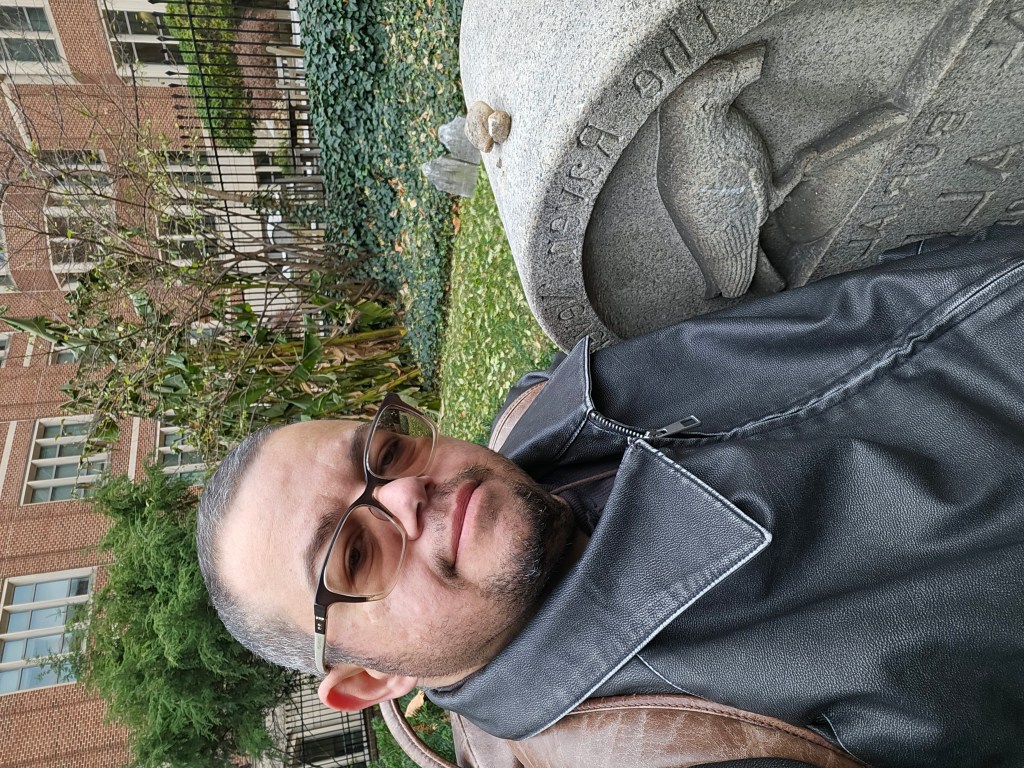 Aim Comperatore wearing glasses and a backpack sitting next to the gravestone at the original burial place of Edgar Allan Poe.