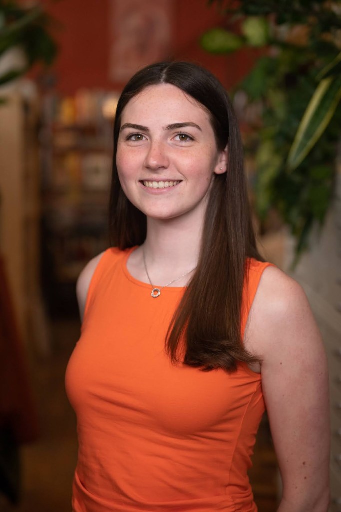 A person with long brown hair stands in front of a bookshelf and indoor plant and they are wearing a orange shirt.