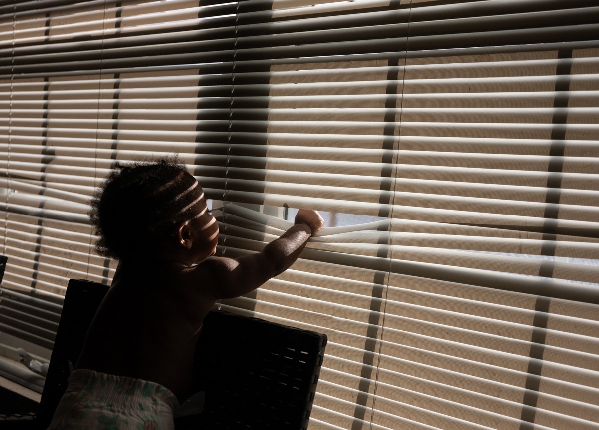 A baby in a diaper puts its fist through a set of blinds to look outside as strips of light fall on its face.