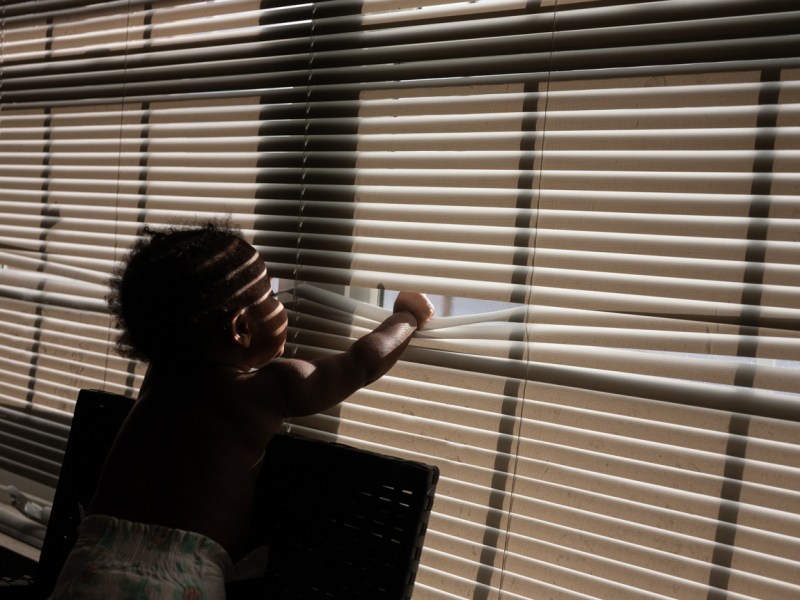 A baby in a diaper puts its fist through a set of blinds to look outside as strips of light fall on its face.
