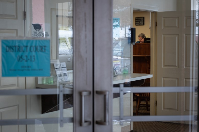 Magisterial District Judge Eugene Riazzi is seen through a series of doors as he hears a landlord/tenant case in his courtroom. "THIS OFFICE HAS 24-HOUR CAMERA SURVEILLANCE" reads a sign on the wall beside the court service window. A container of hand sanitizer sits amongst brochures for related court information on a ledge.