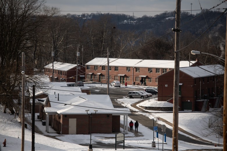 Snow lines the hillside around Crawford Village Housing Complex as people walk through the parking lot and along a shoveled path. Signs for a bus stop and a pole holding security cameras are in the foreground. In the distance, the blue hills of neighboring Duquesne.