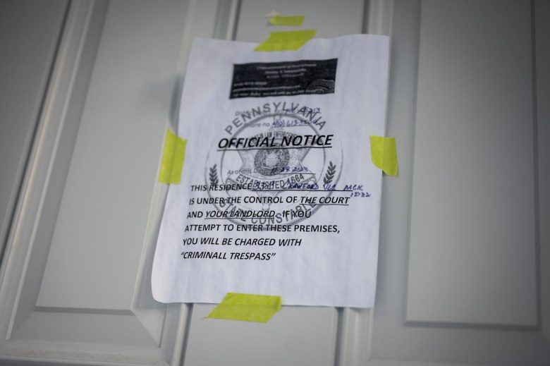 "OFFICIAL NOTICE" reads the black ink of an eviction notice taped to a white front door with yellow tape in McKeesport. A hand-written date, court phone number, and address is added in marker. "IF YOU ATTEMPT TO ENTER THESE PREMISES, YOU WILL BE CHARGED WITH "CRIMINALL TRESPASS" reads the bottom of the page in all-capital letters in front of a law enforcement seal.