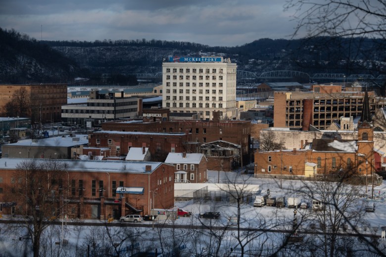 "Discover McKeesport" reads a red, white, and blue sign above the industrial city's downtown district. A blue bridge crosses the Monongahela River in the background. Snow sits on the town roofs and streets.