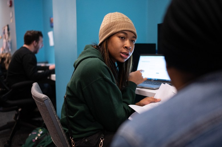 London Reese-Scaife, a housing support clerk, points towards her computer as she holds paperwork while talking with a person facing eviction at the Housing Stabilization Center, Thursday, Jan. 18, 2024, in downtown Pittsburgh. Reese-Scaife wears a beanie and sweatshirt, the center walls are blue. (Photo by Stephanie Strasburg/PublicSource)