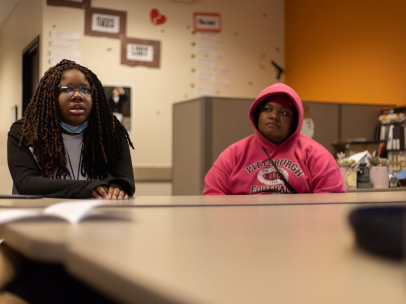 Takara Pack (left) and Sarah Nervais share their thoughts about the root causes of gun violence and ways to prevent shootings while sitting at Youth Enrichment Services' Office in East Liberty on Dec. 7. Behind them: A wall of "Loved Ones Lost," including the names of nine teens the organization has lost to gun violence in recent years, including its own members and their friends and relatives. (Photo by Benjamin Brady/PublicSource)