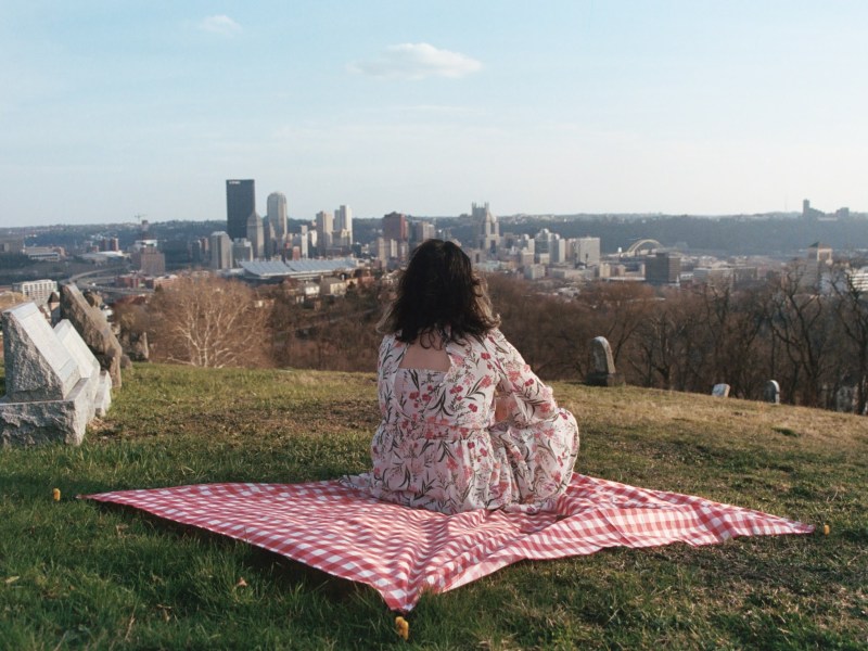 Talia Paone, 27, sits on a red and white gingham blanket in St. John’s Lutheran Cemetery in the Spring Hill–City View neighborhood on April 4, 2023. Since the beginning stages of her transition as a transgender woman, Talia frequently drove up to the cemetery to look at the view of the Downtown skyline and ponder about life — and death.