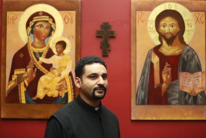 Rev. Paul Abernathy, priest at St. Moses the Black Orthodox Church in the Hill District and CEO of the Neighborhood Resilience Project, stands in front of a red wall with religious images behind him. (Photo by Ryan Loew/PublicSource)