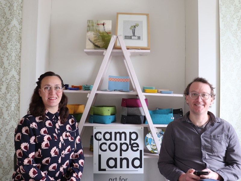Alyssa Kail, a white woman wearing glasses, and Drew Kail, a white man wearing glasses pose in the Camp Copeland studio in front of a display of home goods and a Camp Copeland sign.