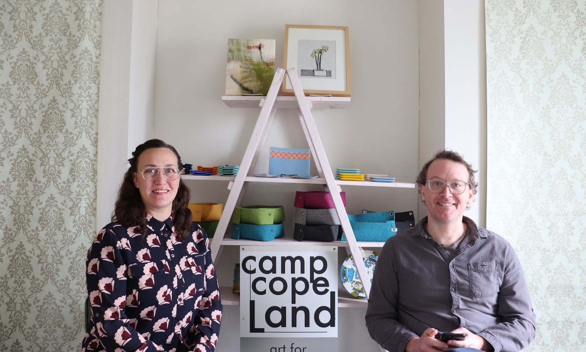 Alyssa Kail, a white woman wearing glasses, and Drew Kail, a white man wearing glasses pose in the Camp Copeland studio in front of a display of home goods and a Camp Copeland sign.