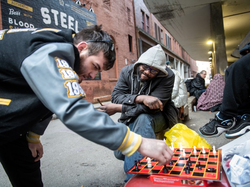 Lucas, left, in a football jacket, and Jamar, center, in a hoodie, play chess atop Jamar's grill together in an alley across from the Smithfield United Church of Christ shelter on Thursday, March 2, 2023, in Downtown. As the shelter closes at 7 a.m., Jamar sweeps the alley and sets up the chessboard he bought at the neighboring toy store, S.W. Randall Toyes & Gifts. (Photo by Stephanie Strasburg/PublicSource)