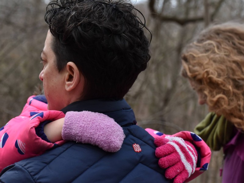 Lee Brigido carries his foster child in his arms as he walks down a trail next to Mary Carroll in Frick Park on Saturday, March 18, 2023. When there’s cause to advocate for his foster kid, “I put my fighting gloves on a lot faster,” Brigido said, laughing. (Photo by Amaya Lobato-Rivas/PublicSource)