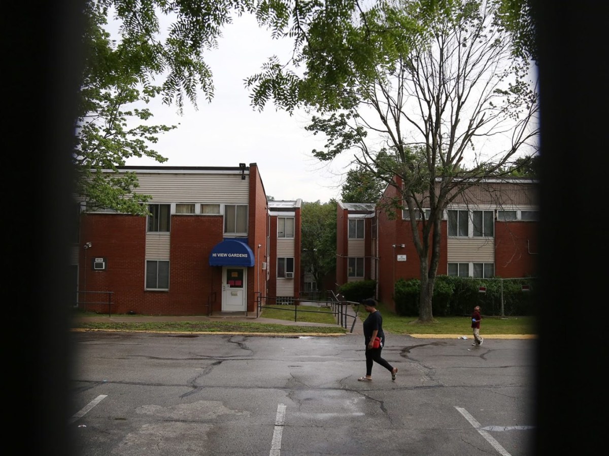 A resident of Hi View Gardens and her young son, seen through a gate, cross the main parking lot of the five-building McKeesport complex. The Allegheny County Health Department found 347 housing health code violations at Hi View and nearby Midtown Plaza from 2017 through 2020, and it collected $2,500 in penalties. (Photo by Ryan Loew/PublicSource)