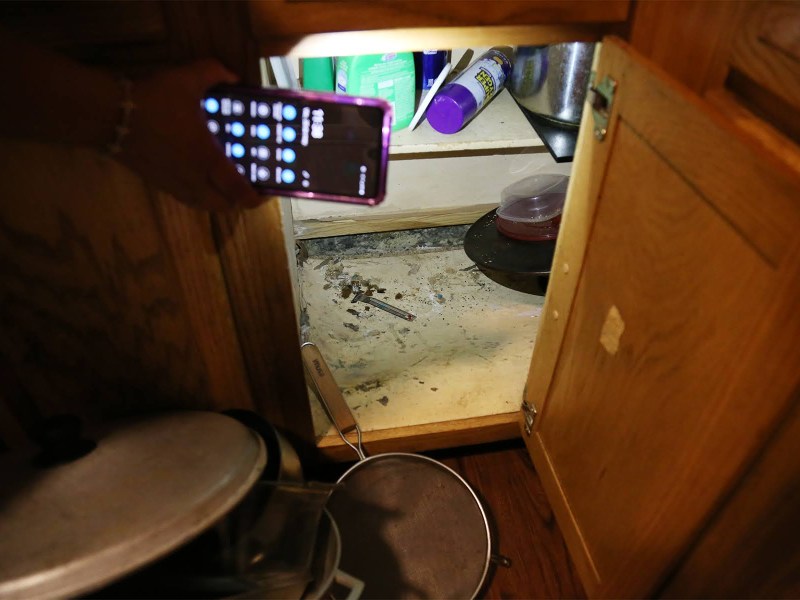 Beatrice Román, a tenant of Hi View Gardens whose former apartment was inspected by the Allegheny County Health Department four times this year — for mice and roaches, leaks, no heat and holes in wall — uses her phone's flashlight to show the inside of a kitchen cabinet. (Photo by Ryan Loew/PublicSource)