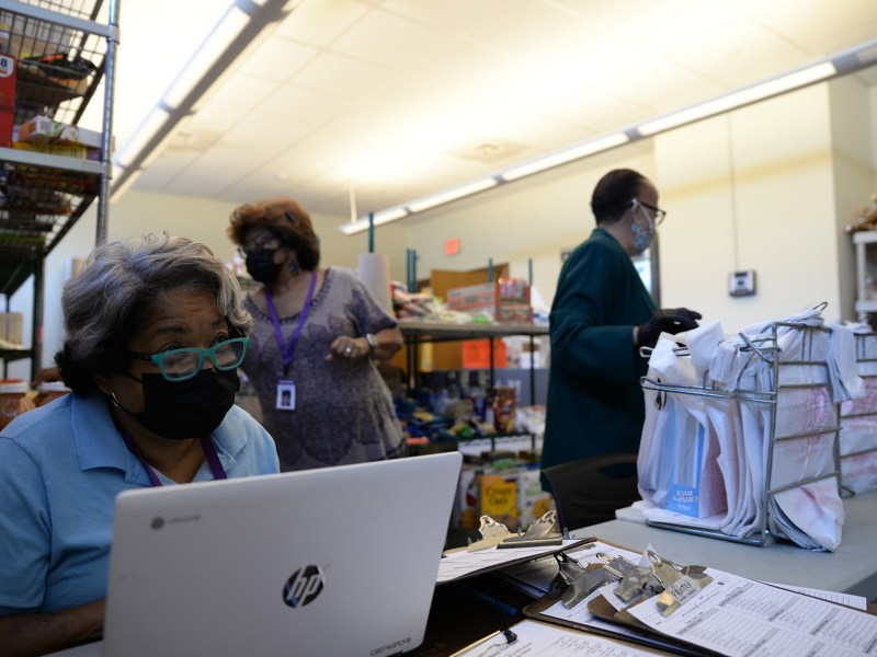 A volunteer sits in front of a computer at the lower left side of the photo, with two other volunteers moving behind them. There are shelves of food in the background.