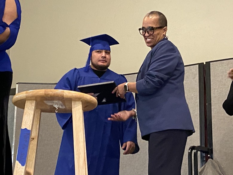 A man in a blue graduation gown receives a certificate from a woman in a blue suit next to a podium.