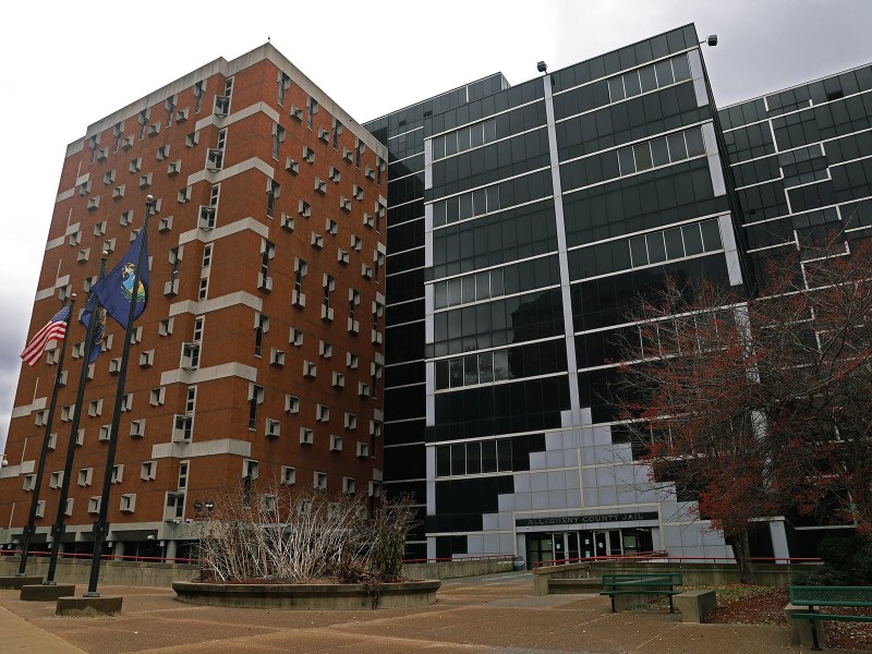 The front facade of Allegheny County Jail. The building is constructed of long, rectangular glass panels in the center and red brick on the left side.