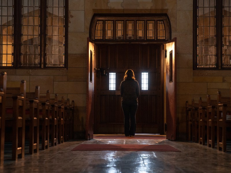 Jane, who wishes to remain anonymous, stands silhouetted in the doorway of a church with pews on either side.