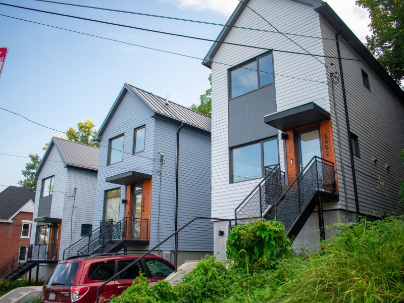 Three tall, narrow homes with gray siding, black roofs and wooden accents by the front doors sit side by side. A red car is parked in the driveway of the rightmost home. Each home has a mix of concrete and metal stairs leading up to the front door.