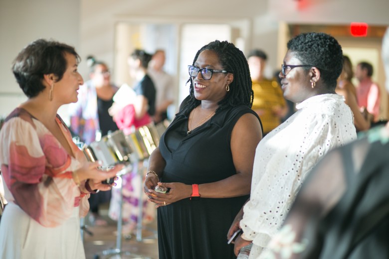 Ascender CEO Nadyli Nuñez stands to the left of Joyce Howard and her daughter, and uses her hands while speaking. The women are talking in a room with other people. 