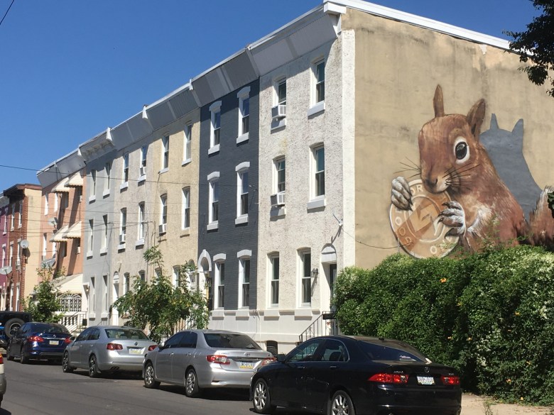 Patricia De Carlo’s Philadelphia neighborhood of Norris Square in summer 2023. Photo shows a squirrel mural on the side of neutral colored stucco row homes. Cars parked on the street. (Courtesy of Patricia De Carlo)