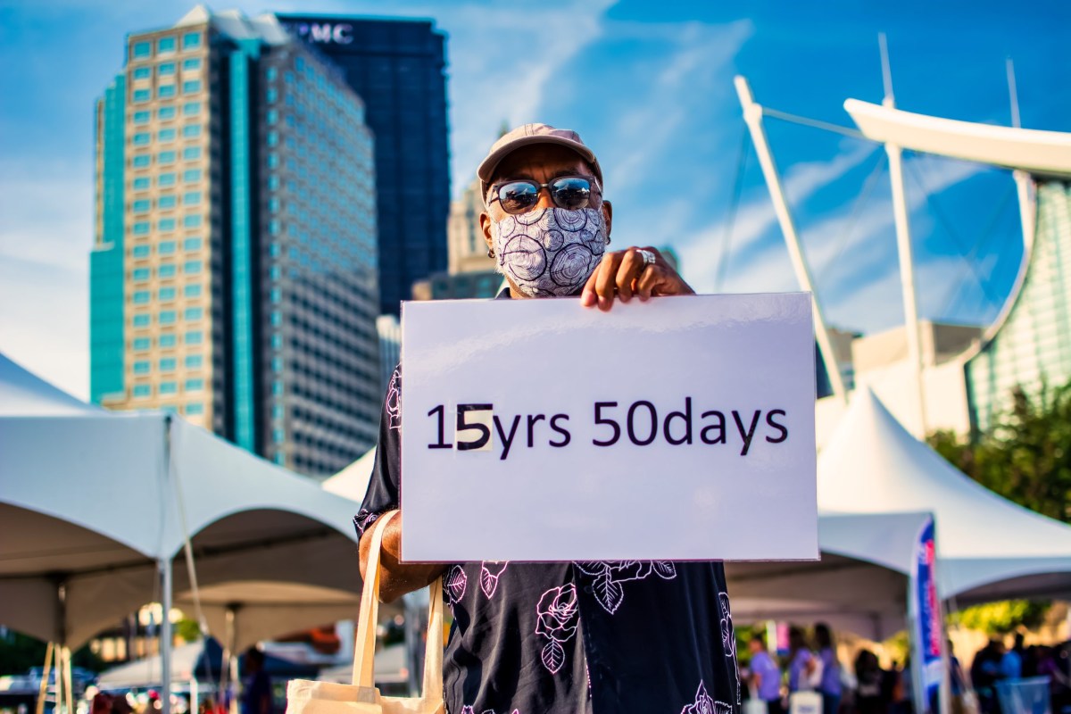 A person wearing sunglasses and mask holds a sign reading "15 years 50 days" in front of the Pittsburgh skyline