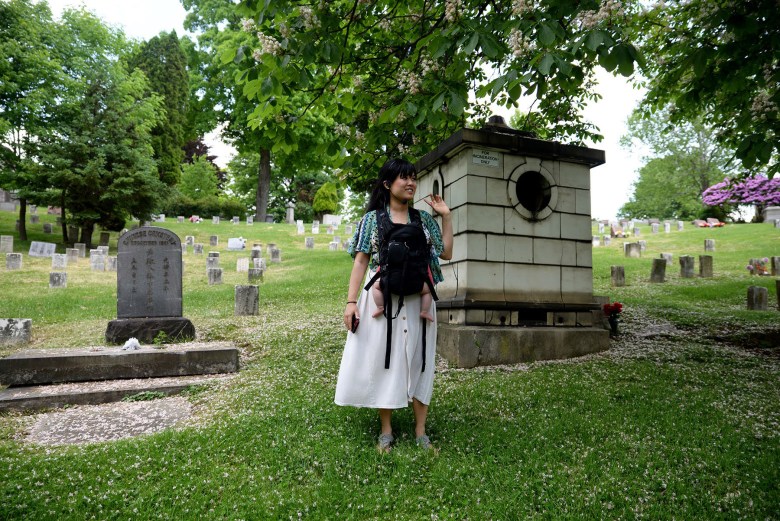 Lena Chen stands on grass in the Chinese Cemetery holding a baby in a baby carrier.