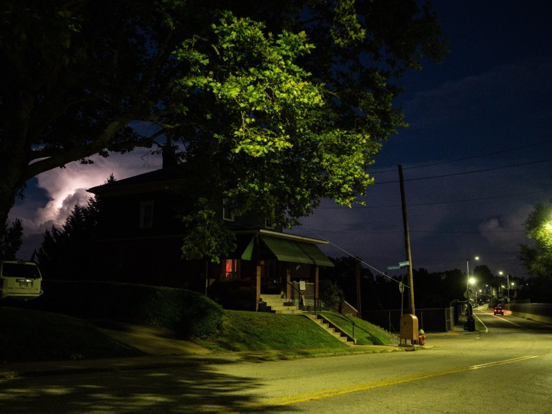 Lightning lights up a cloud beyond the 3400 block of Shadeland Avenue on Saturday, Aug. 6, 2022, in Brighton Heights. A house is partially covered by a tree lit by a streetlight, which casts a green tint on the street below, stretching over a bridge in the distance. A ShotSpotter acoustic sensor alerted Pittsburgh police to sounds, interpreted as gunshots, around 3400 Shadeland Avenue in Brighton Heights on Dec. 15, 2018. That data was used in the prosecution of Angelo Weeden, who is now appealing his conviction to the Pennsylvania Supreme Court, in a case that may have implications for the use of gunshot detection technology statewide and beyond. (Photo by Stephanie Strasburg/Public Source)