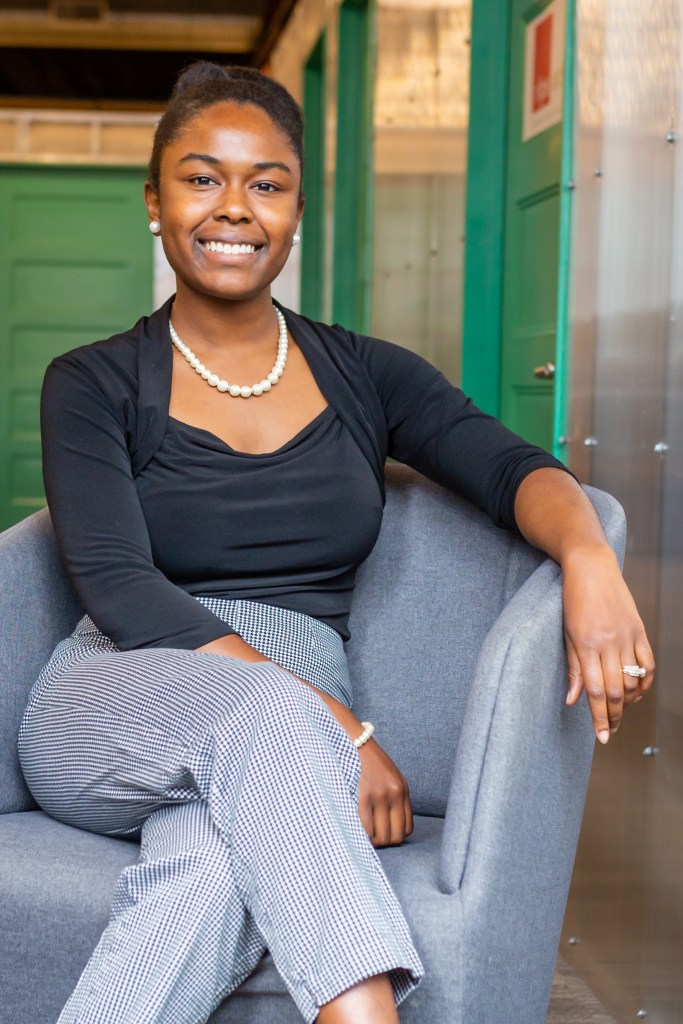 Talpha Harris, a Black woman wearing a black blouse and pearl necklace, sits in a grey chair with her legs crossed. 